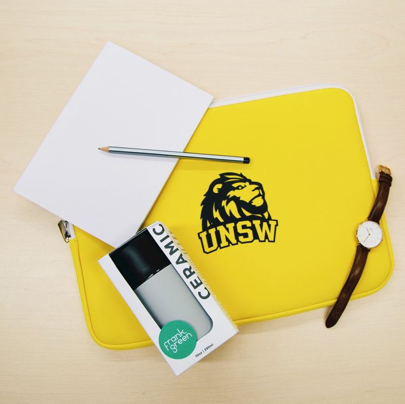 Bright yellow laptop sleeve with lion and UNSW logo in black. Laid out with a notebook, pencil, watch and frank green keep cup.