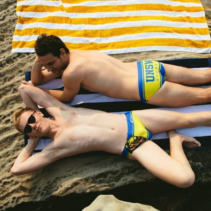 Two men wearing UNSW budgie smugglers on a towel on the sand.
